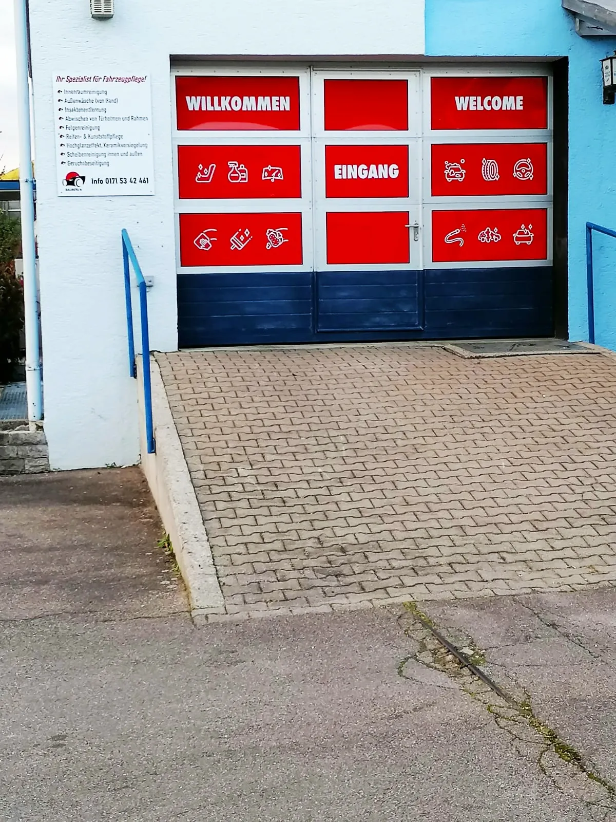 Einfahrt von Sauberle Fahrzeugpflege mit rotem Hallentor und der Leistungstafel an der Wand